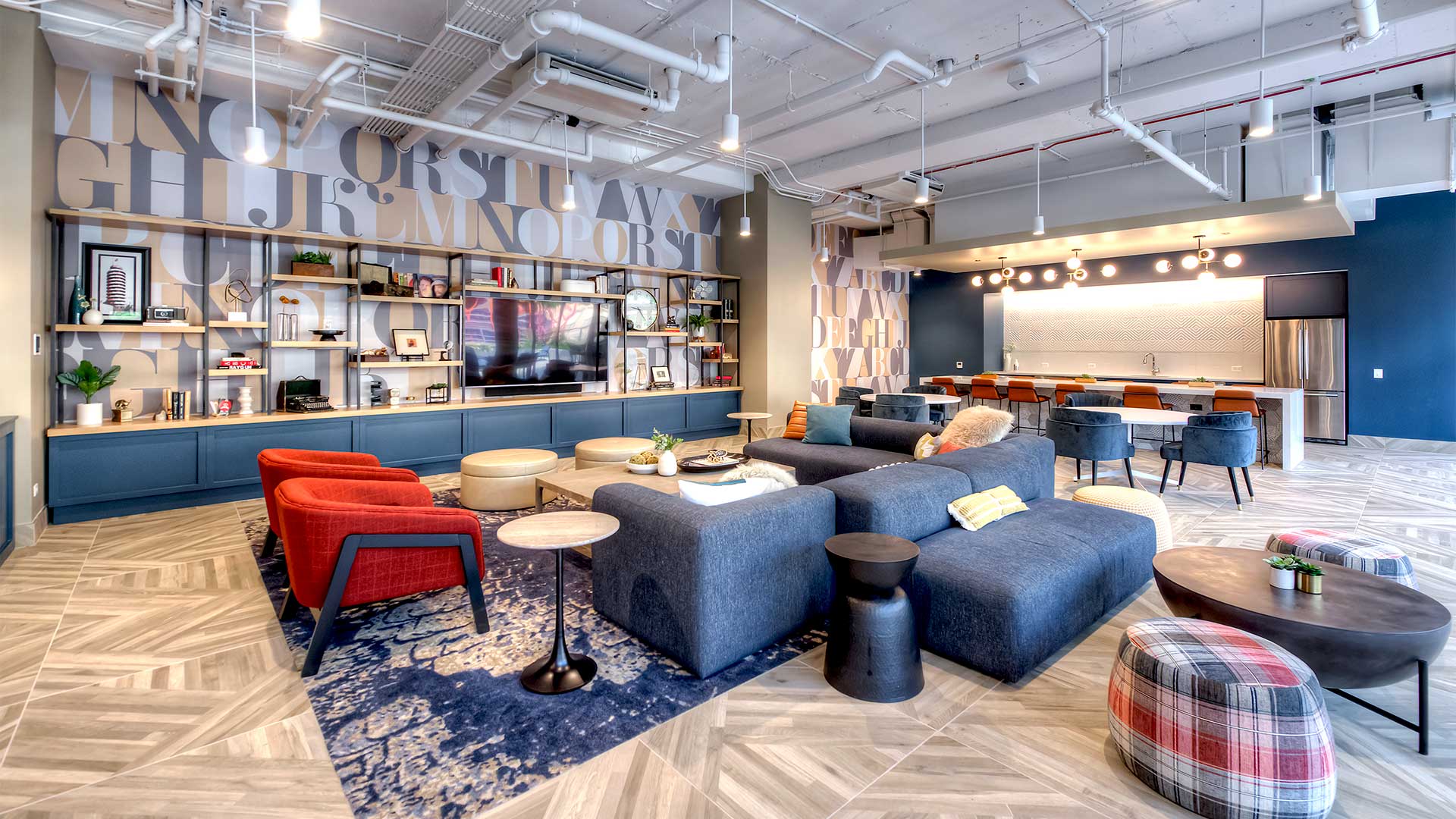 A collection of comfortable chairs and sofas sits in the middle of the room. The back wall to the left is a large bookshelf with a variety of objects on it and a large television. The back wall to the right is a coffee bar.