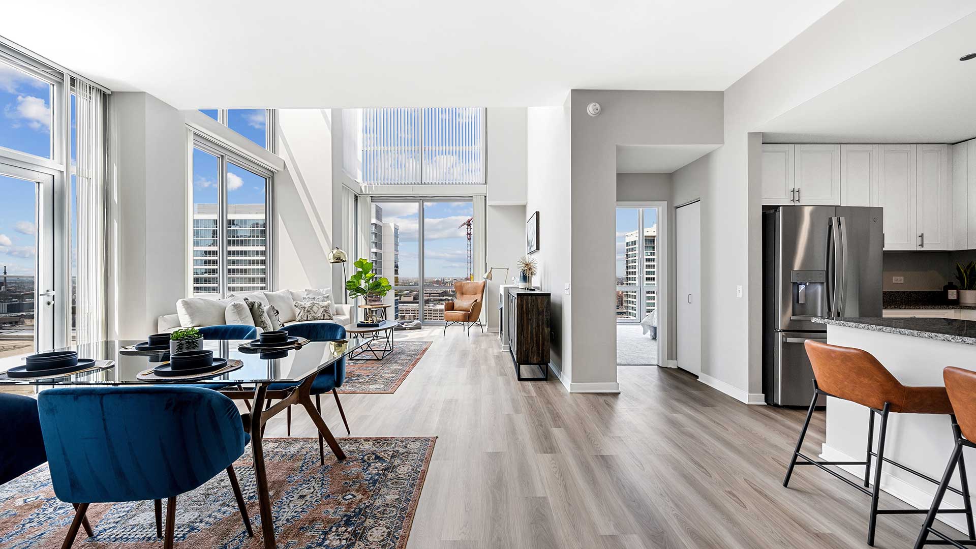 A living room in a residence at Burnham Pointe. The room is long with windows reaching high up out of view. To the left is a dining set with a living room set behind. To the right is the kitchen with two bar stools sitting before the counter.