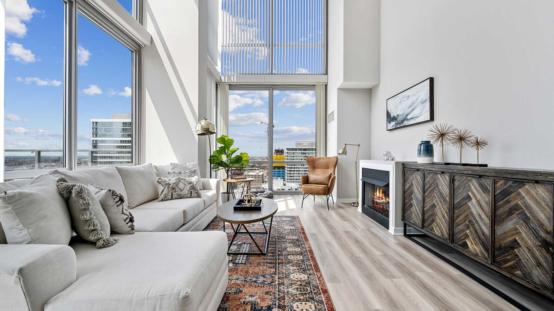 A long living room with windows that rise high up out of the image. A couch runs in front of the windows on the left and a dresser and fireplace line the right wall.