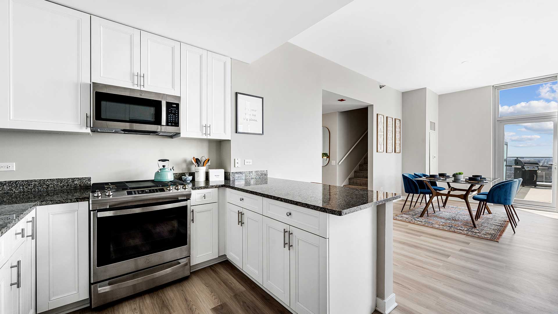 Standing in a kitchen in a residence at Burnham Pointe. The oven and microwave are seen on the left with white cabinets all around. A counter is in the middle separating the kitchen from the dining area beyond.