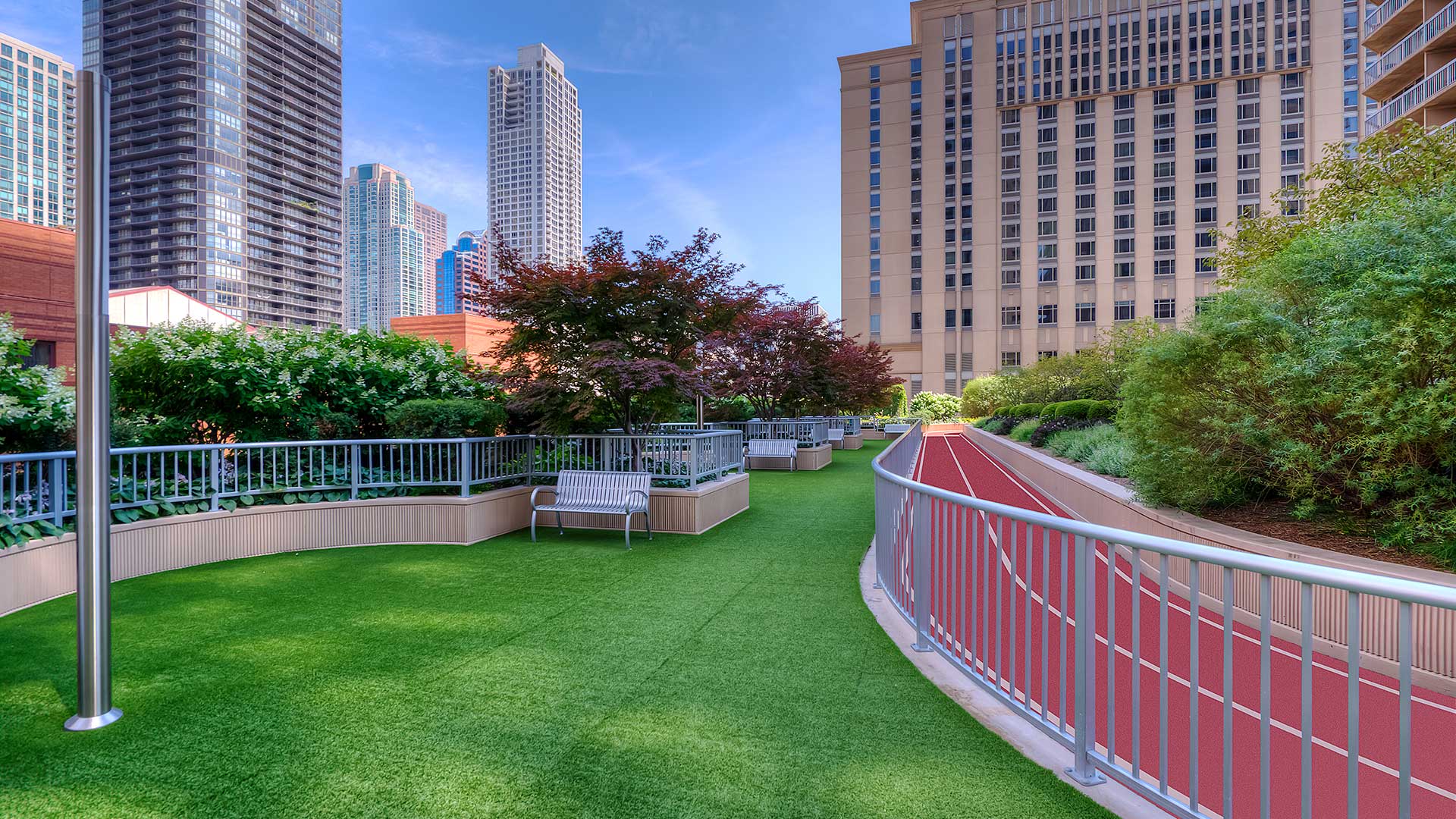 An outdoor dog run with green turf. It is fenced in with benches and foliage on the left and an outdoor running track on the right. City buildings are seen in the background.