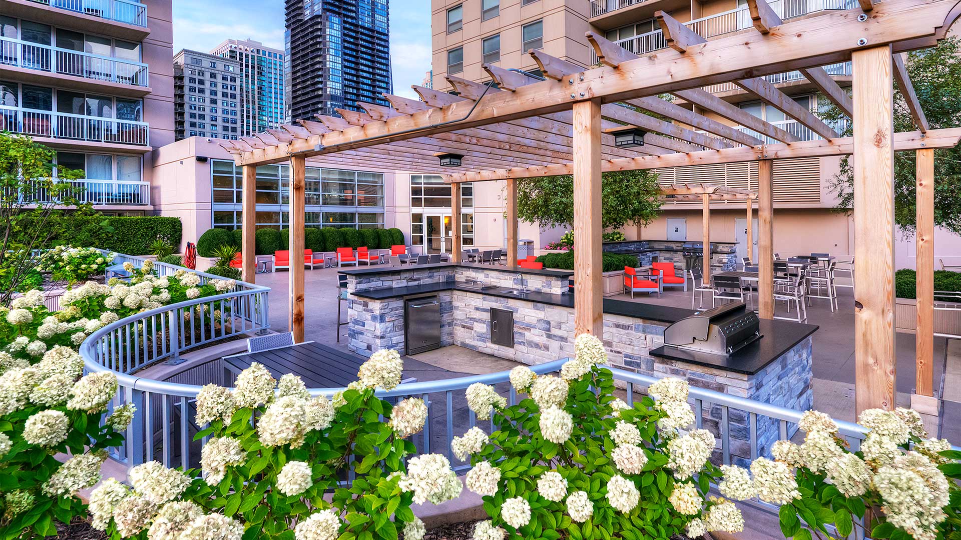 Looking over a row of white flowers into the outdoor kitchen area. A grilling station is close by under a pergola and there is seating beyond.