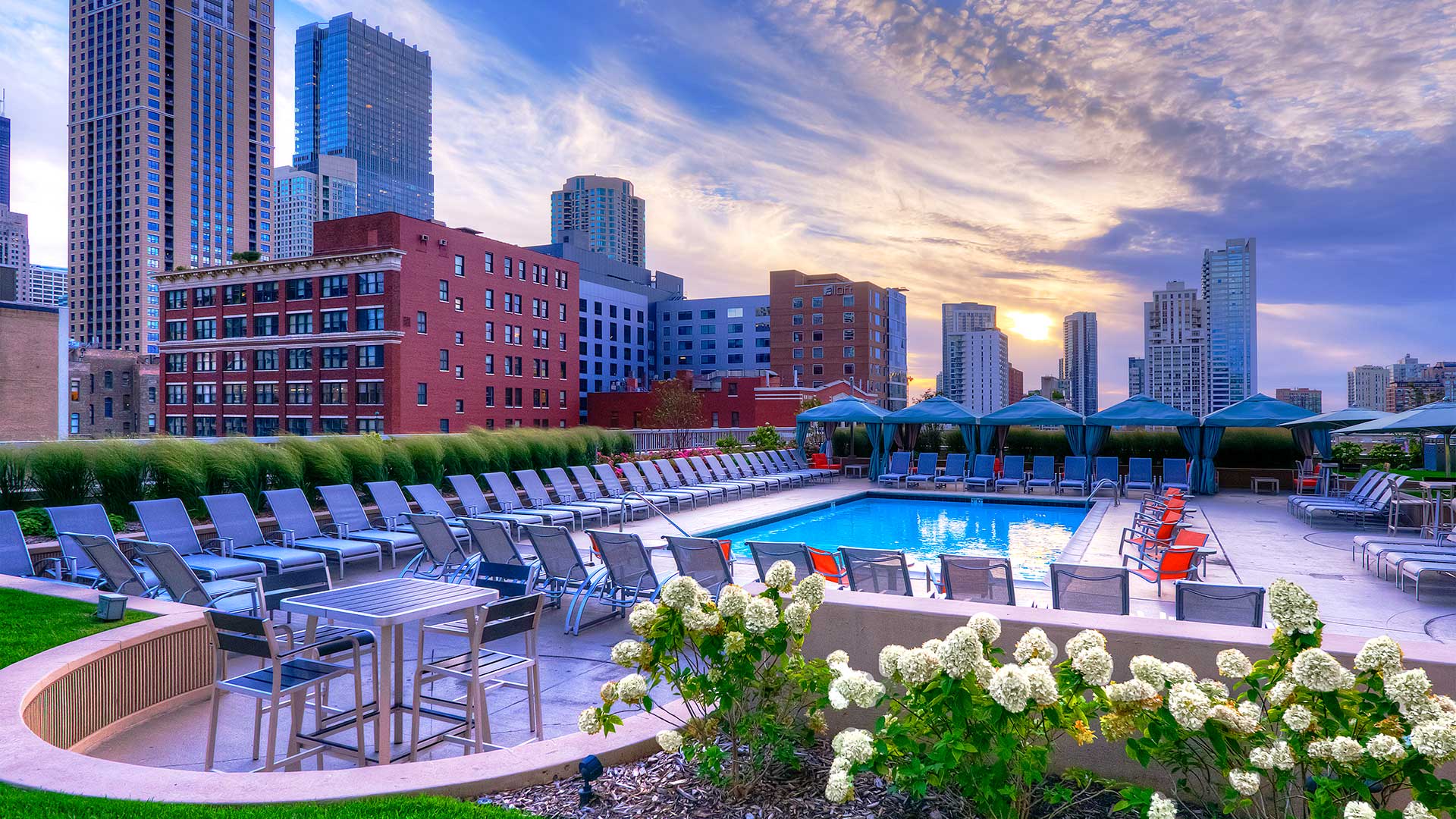 Looking over a row of white floors at the pool deck at dusk. The sun is setting ahead and there are city building all around.
