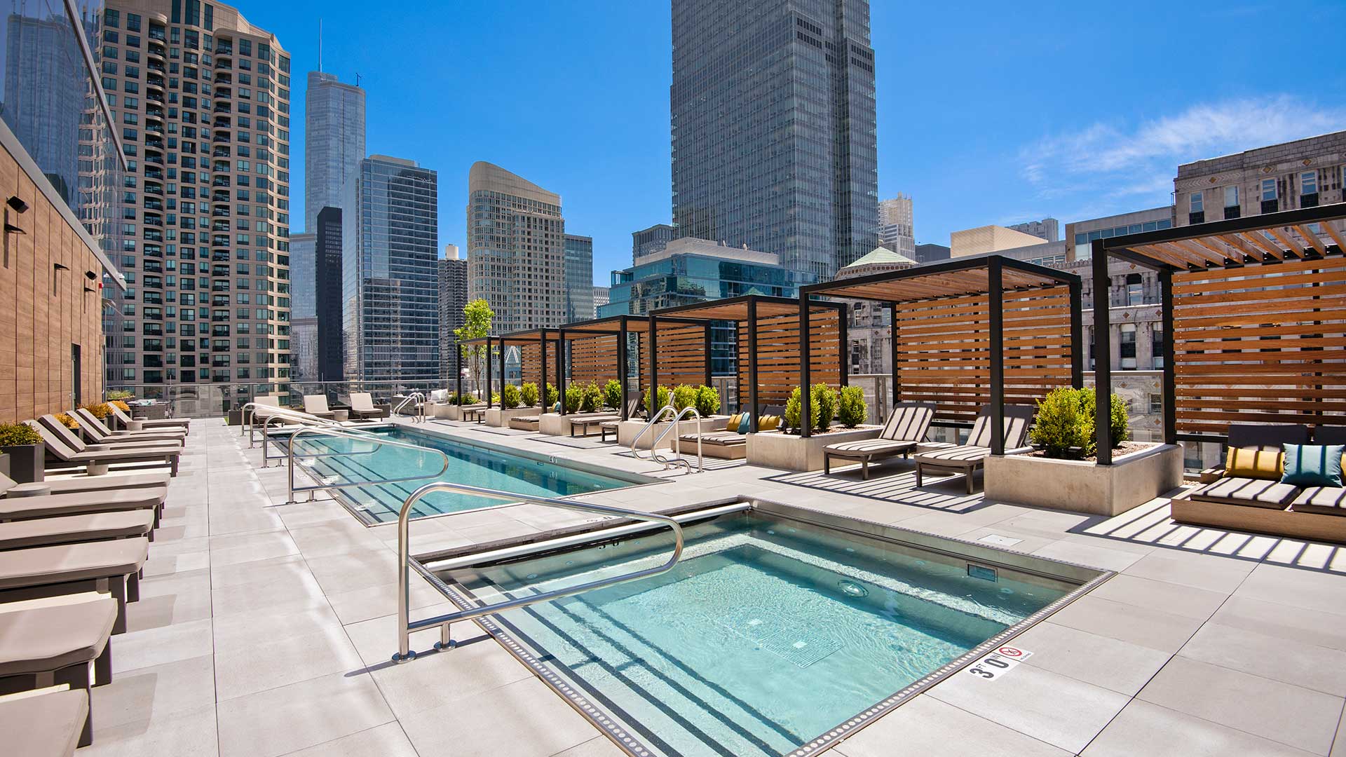 Looking across the outdoor hot tub and then the pool on the amenity deck. A row of pergolas and lounge chairs lines the far side. City buildings are seen rising in the background.