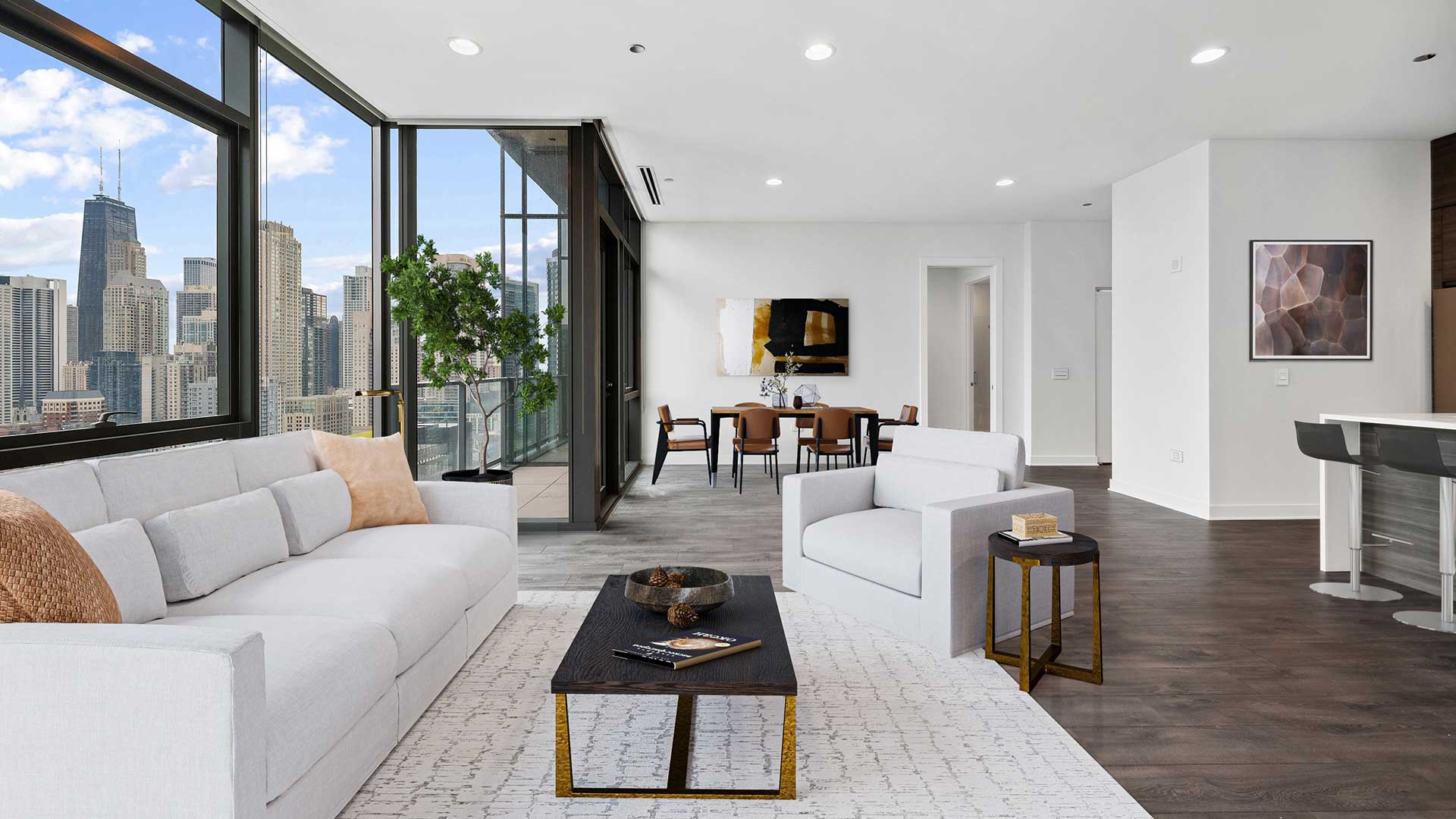 A large living room in a Hubbard221 residence. There is a living room set in the foreground with a couch, chair and coffee table. Beyond is a dining set. The left are floor-to-ceiling windows that show the Chicago skyline. To the right is the kitchen.